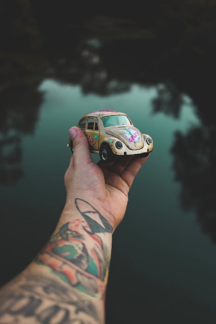 A tattooed hand holds a colorful Volkswagen Beetle toy car against a serene lake backdrop.
