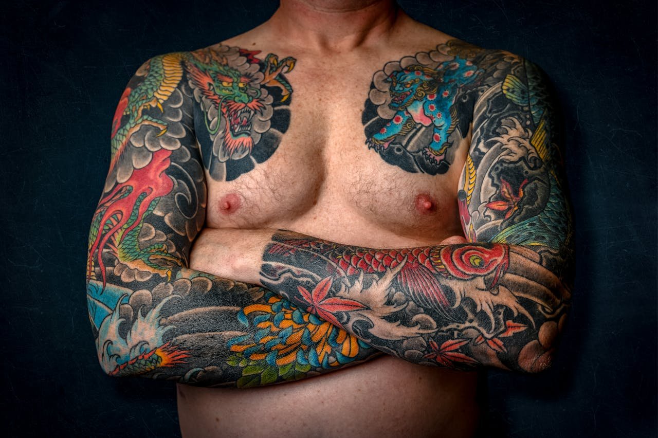 Close-up of a shirtless man with colorful sleeve tattoos and chest art.