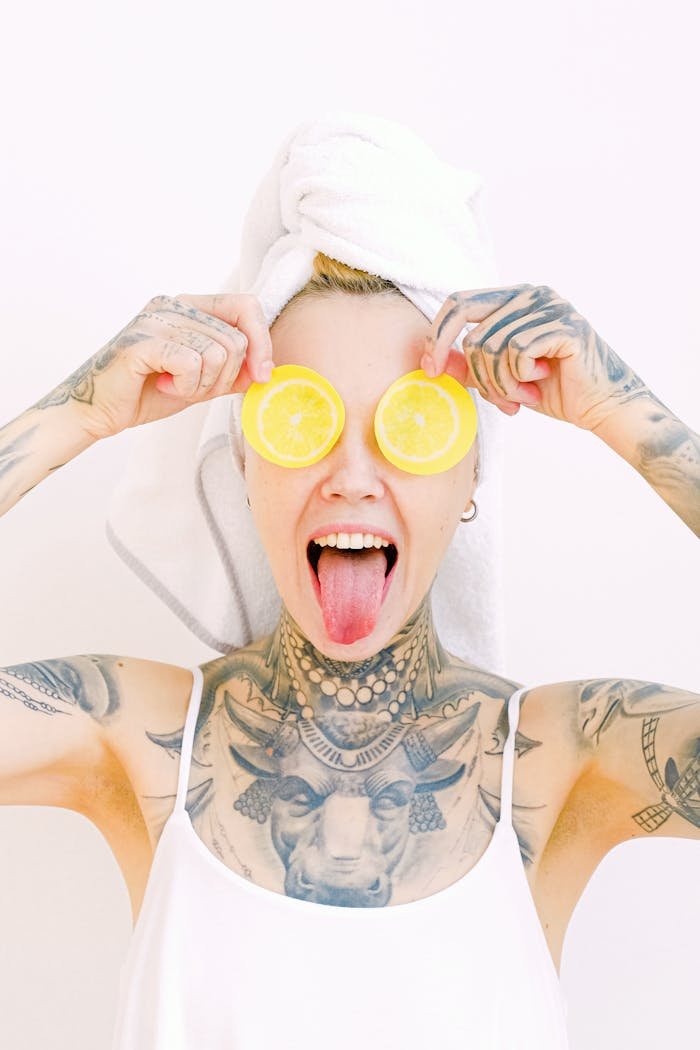 Fun portrait of a tattooed woman with towel turban, playfully holding lemon slices and sticking out her tongue.