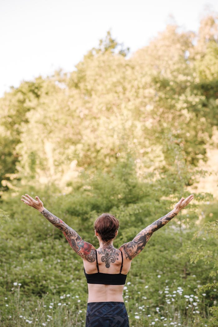 Back view of a tattooed woman practicing yoga in a sunny outdoor setting, arms raised for wellness and vitality.